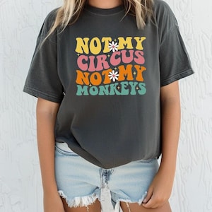 May include: A dark gray t-shirt with a retro-style graphic that reads "Not My Circus Not My Monkeys" in colorful letters with flowers.