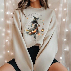 May include: A beige crewneck sweatshirt featuring a graphic illustration of a witch wearing a hat and holding a glowing lantern. The design includes scattered autumn leaves, small bats, and the text "If you've got it haunt it" in black.