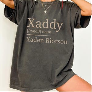 May include: Dark gray t-shirt with the word "Xaddy" in a light brown font. Below the word is the phonetic pronunciation and the word "noun". Underneath is the name "Xaden Riorson".