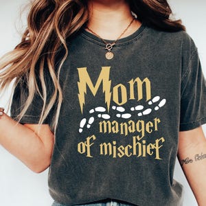 May include: A dark gray t-shirt with a gold lightning bolt and the text "Mom manager of mischief" in a gold font. The text is surrounded by white footprints.