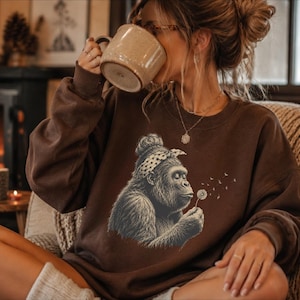 May include: A dark brown long-sleeve crewneck sweatshirt featuring a graphic print of a gorilla wearing a polka-dot headband and blowing on a dandelion. The illustration is rendered in shades of gray and white, centered on the chest of the garment.