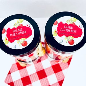 May include: Two glass jars with black lids, filled with play dough and pizza-themed accessories. Each jar has a label that reads "CASA BLEU PIZZA PLAY DOUGH" with a tomato and pizza slice design. The jars sit on a red and white checkered cloth.