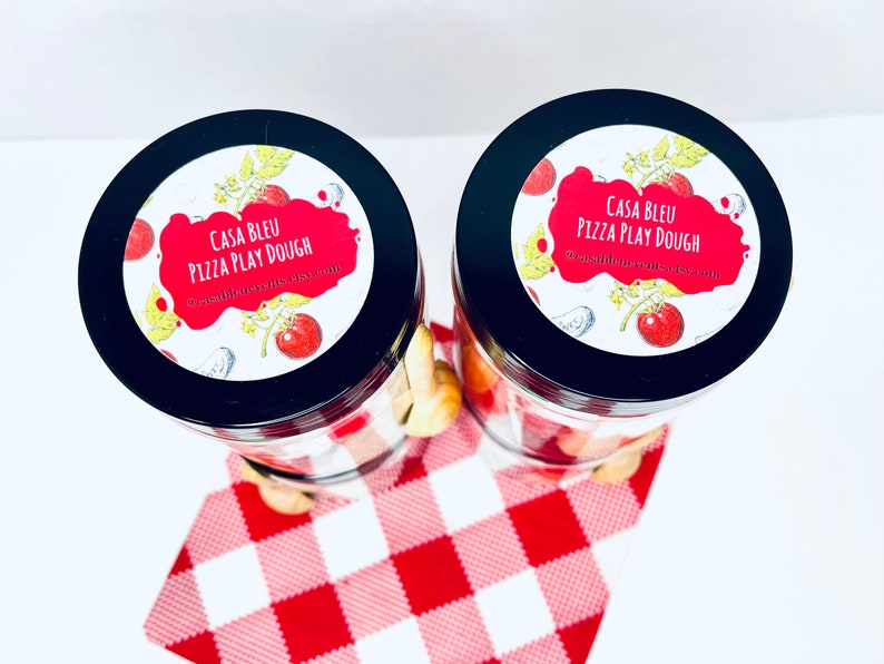 May include: Two glass jars with black lids, filled with play dough and pizza-themed accessories. Each jar has a label that reads "CASA BLEU PIZZA PLAY DOUGH" with a tomato and pizza slice design. The jars sit on a red and white checkered cloth.