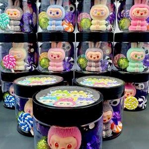 Könnte beinhalten: Eine Sammlung durchsichtiger Kunststoffgläser mit schwarzen Deckeln, die jeweils eine kleine Plüschhasenfigur in verschiedenen Pastellfarben sowie bunte Wirbellollis und glitzernde Kugeln enthalten. Die Deckel sind mit einer Grafik und dem Text Happy Halloween from Addta versehen.