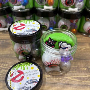 Könnte beinhalten: Transparente Kunststoffdosen gefüllt mit hellgrünem Schleim, darin enthalten sind Kunststoff-Augäpfel, kleine Knochenanhänger, glitzernde Kugeln und Spielfiguren. Die schwarzen Deckel zeigen ein Ghostbusters-Motiv mit dem Text Happy Birthday Ben und I ain't afraid of no ghosts.