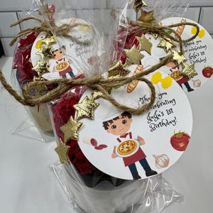 May include: Three party favors wrapped in clear cellophane. Each favor is decorated with a round tag featuring a cartoon chef holding a pizza. The tags are adorned with gold star embellishments and tied with twine. Text on the tag reads "Thank you for celebrating JuJu's 1st Birthday!"