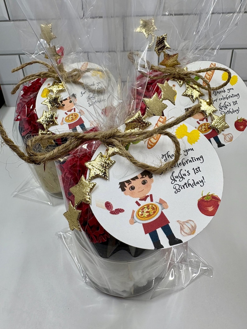 May include: Three party favors wrapped in clear cellophane. Each favor is decorated with a round tag featuring a cartoon chef holding a pizza. The tags are adorned with gold star embellishments and tied with twine. Text on the tag reads "Thank you for celebrating JuJu's 1st Birthday!"