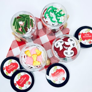 May include: Four clear jars filled with play dough designed to resemble pizza toppings, arranged on a red and white checkered cloth. The jars are surrounded by black-lidded containers with the text "CASA BLEU PIZZA PLAY DOUGH".