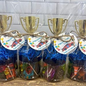 K&ouml;nnte beinhalten: Vier durchsichtige Kunststoffgl&auml;ser gef&uuml;llt mit blauem Papierstreifen und Spielzeug-Rennwagen, gekr&ouml;nt von goldenen Kunststoff-Pokalen. Jedes Glas hat ein rundes Etikett, das mit einer Schnur befestigt ist und Happy birthday guy zeigt.