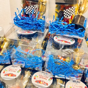 K&ouml;nnte beinhalten: Transparente Kunststoff-Geschenkboxen gef&uuml;llt mit blauem Papierstreifen, einem kleinen goldenen Kunststoffpokal, einer Zielflagge und verschiedenen kleinen Rennsport-Artikeln. Jeder Deckel hat einen runden Aufkleber mit rotem Rennwagen und Text.