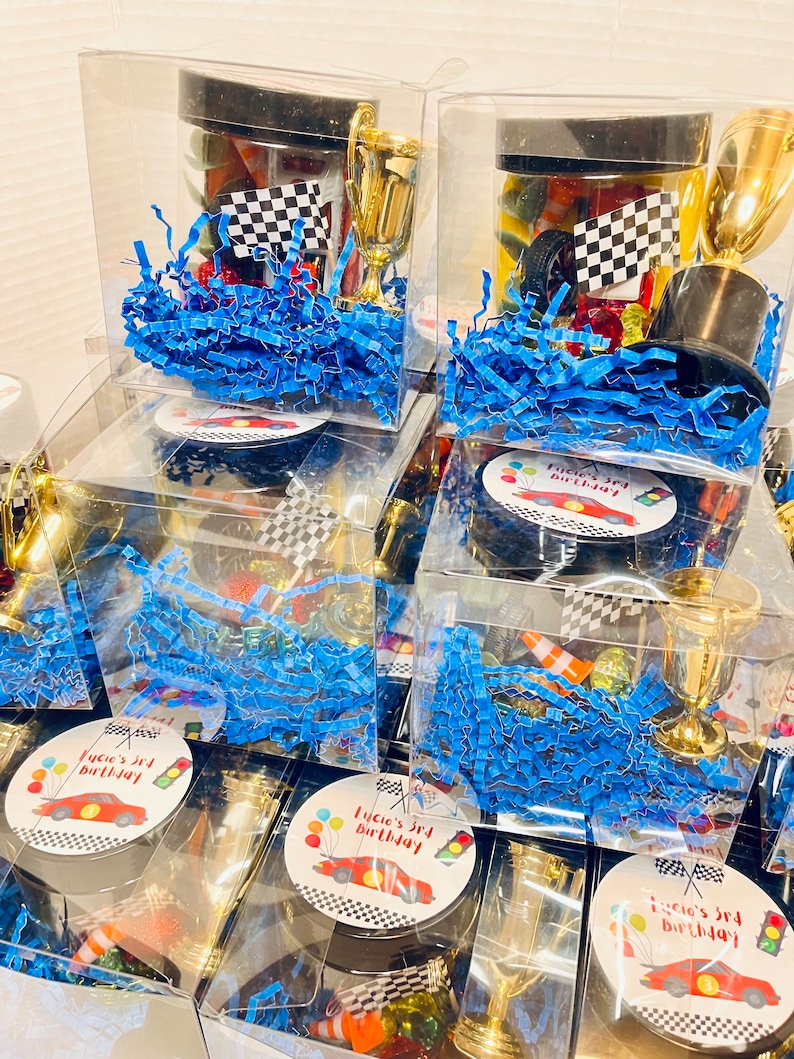 K&ouml;nnte beinhalten: Transparente Kunststoff-Geschenkboxen gef&uuml;llt mit blauem Papierstreifen, einem kleinen goldenen Kunststoffpokal, einer Zielflagge und verschiedenen kleinen Rennsport-Artikeln. Jeder Deckel hat einen runden Aufkleber mit rotem Rennwagen und Text.