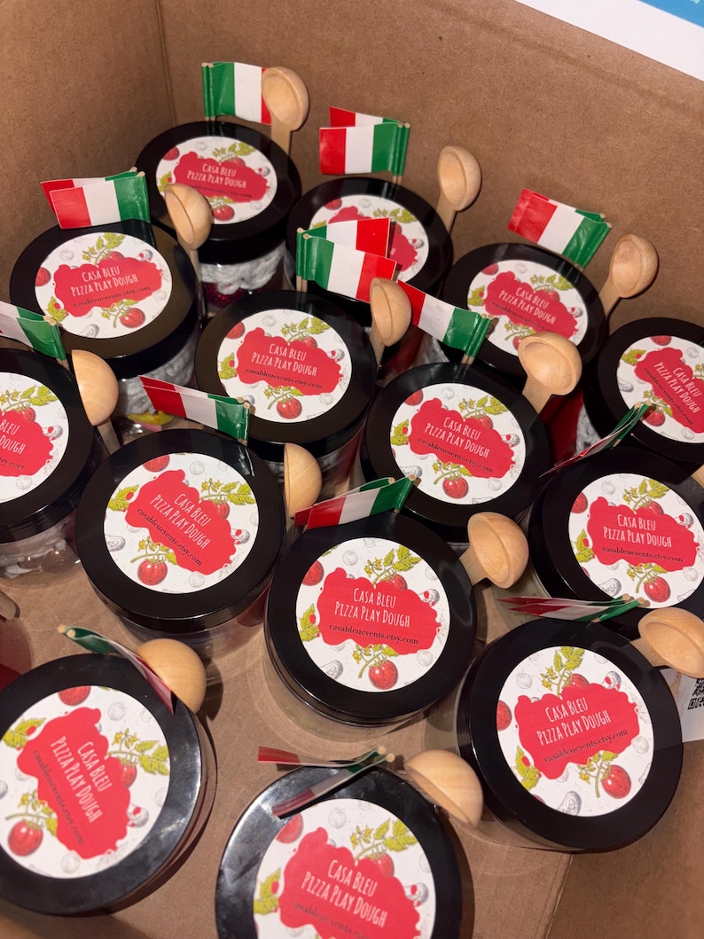 May include: A selection of black-lidded jars, each bearing a label reading "Casa Bleu Pizza Play Dough", accompanied by a small wooden spoon and a miniature Italian flag. The jars are presented in a cardboard box.