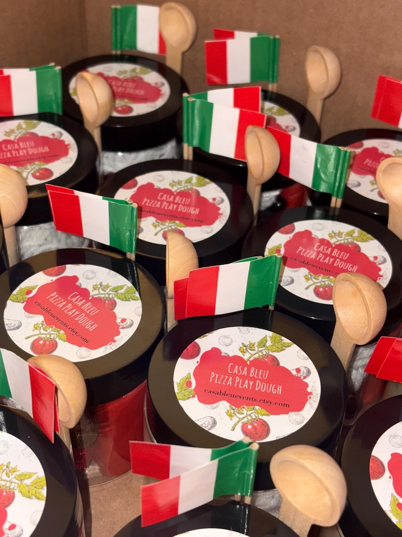 May include: A collection of Casa Bleu Pizza Play Dough jars, each with a label showing tomatoes and the text "Pizza Play Dough." Small Italian flags and wooden spoons are arranged with the jars. The website casableuevents.etsy.com is also visible.