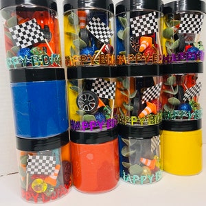 K&ouml;nnte beinhalten: Eine Sammlung durchsichtiger Kunststoffgl&auml;ser mit schwarzen Deckeln, gef&uuml;llt mit farbigem Sensorknete und Rennsport-Zubeh&ouml;r wie Miniatur-Zielflaggen, orangefarbenen Verkehrskegeln, Spielzeugautoreifen und dem Schriftzug HAPPY BDAY in Metallbuchstaben.