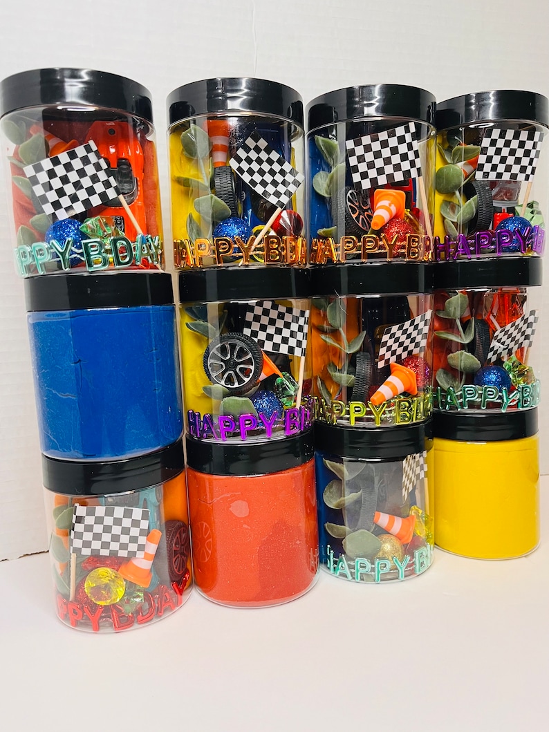 K&ouml;nnte beinhalten: Eine Sammlung durchsichtiger Kunststoffgl&auml;ser mit schwarzen Deckeln, gef&uuml;llt mit farbigem Sensorknete und Rennsport-Zubeh&ouml;r wie Miniatur-Zielflaggen, orangefarbenen Verkehrskegeln, Spielzeugautoreifen und dem Schriftzug HAPPY BDAY in Metallbuchstaben.