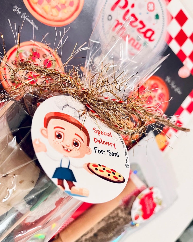 May include: A cellophane-wrapped gift basket with a pizza theme. The basket includes a pizza menu, a tag with a cartoon chef, and the text "Special Delivery For: Soni". The basket is decorated with gold and red tinsel.