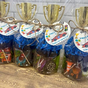 K&ouml;nnte beinhalten: Vier durchsichtige Kunststoffbeh&auml;lter gef&uuml;llt mit blauem Papierstreifen und kleinen Spielzeug-Rennwagen. Jeder Beh&auml;lter ist mit einem goldenen Kunststoffpokal und einem runden Etikett mit rotem Rennwagen und Happy Birthday versehen.