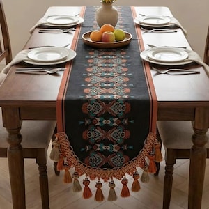 Op de afbeelding: Een eettafel gedekt voor een maaltijd, met een donkere loper met een ingewikkeld patroon in teal, oranje en crème. De loper is versierd met kwastjes. De tafel is gedekt met witte borden, bestek en een vaas met zonnebloemen.