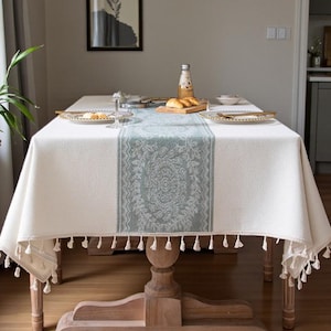 May include: A white rectangular tablecloth with a light blue patterned runner and tassel trim. The tablecloth covers a wooden table with a central pedestal base. The table is set with plates, glasses, and a bottle.