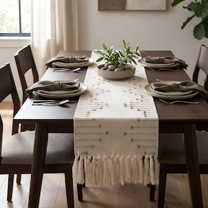 May include: A dining table set for a meal. The dark wood table is set with plates, napkins, and silverware for six people. A cream-coloured table runner with fringe and a decorative bowl with greenery complete the setting.