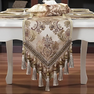 May include: A white table is set with a decorative table runner, placemats, and plates. The runner features a damask pattern in beige and brown, with tassel fringe. A vase of roses sits in the center, enhancing the elegant dining setting.