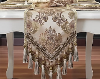Op de afbeelding: Een witte tafel is gedekt met een decoratieve tafelloper, placemats en borden. De loper heeft een damastpatroon in beige en bruin, met kwastjes. Een vaas met rozen staat in het midden, wat de elegante setting verbetert.