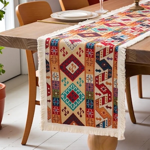 May include: A patterned table runner in shades of red, blue, orange, and beige, with a fringed edge. The runner is displayed on a wooden table. The design incorporates geometric diamond and arrow motifs.