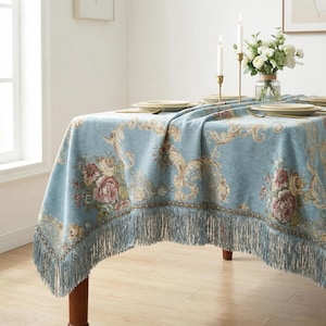Puede incluir: Un mantel azul claro con un estampado floral y flecos. El mantel presenta un diseño detallado con flores rosas y beige y adornos decorativos. Los flecos añaden un toque clásico a la mesa.