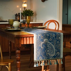 Könnte beinhalten: Blaues Tischläufer mit Paisley-Muster, cremefarbenem Rand und Quasten. Der Läufer liegt auf einem Holztisch mit einem Essarrangement, einschließlich Gläsern, Tellern und einem Kerzenhalter. Der Tisch ist von Holzstühlen umgeben.