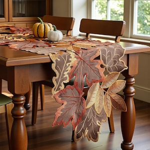 May include: A brown table runner with embroidered autumn leaves in shades of brown, orange, and gold. Two small pumpkins, one orange and one white, sit on the table. The table is made of wood and has a warm, inviting appearance.
