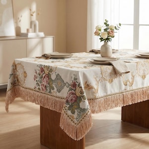 Può includere: Tovaglia color crema con un motivo floreale nei toni del rosa, verde e oro. La tovaglia ha un bordo con frange ed è appoggiata su un tavolo di legno. Un vaso di fiori e delle posate sono visibili.
