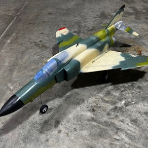 May include: A green, tan, and black model airplane with a red stripe on the tail. The plane has a camouflage paint scheme and is sitting on a gray concrete surface.