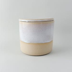 May include: A ceramic jar with a lid. The jar is cylindrical, with a light beige base and a white upper section that fades into a soft pink near the top. The lid matches the top section's colour and shape.