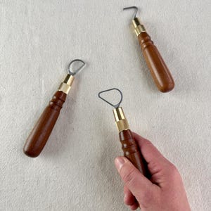 May include: Three pottery loop tools with wooden handles and brass ferrules are arranged on a neutral-coloured surface. One tool is held in a hand. The tools have a curved metal loop or hook at the end, used for shaping and carving clay.