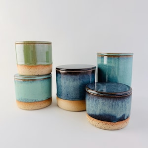 May include: A collection of five cylindrical ceramic canisters with lids. The containers feature a two-tone design with a speckled, unglazed beige base and a smooth, glossy glazed upper section in varying shades of green and deep blue.