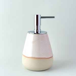May include: A ceramic soap dispenser with a pear-shaped design. The top half is a light pink colour, the bottom half is beige, and the dispenser has a silver pump. The dispenser is designed for use in a bathroom or kitchen.