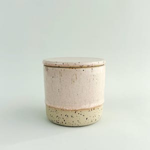 May include: A ceramic jar with a lid. The jar is cylindrical with a light pink glaze on the upper section and a speckled beige base. The lid is the same light pink colour. The jar has a handmade aesthetic.