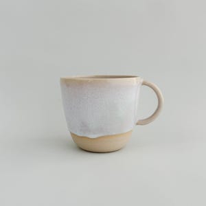 May include: A ceramic mug with a beige base and handle, with a white, textured upper section. The mug has a simple, rounded design, suitable for tea or coffee. The mug is set against a plain, light background.