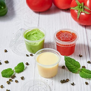 May include: Three clear plastic containers with lids, filled with different coloured sauces. The containers are on a white wooden surface with green leaves and small brown seeds scattered around them.