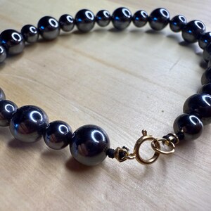 May include: A dark gray beaded necklace with a gold-colored clasp. The beads are spherical and vary in size, reflecting light. The necklace is displayed on a light brown wooden surface.