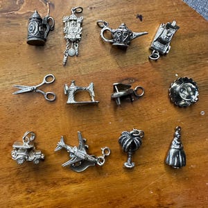 May include: A collection of 12 silver charm pendants, including a sewing machine, a teapot, a pair of scissors, a cuckoo clock, a palm tree, a car, an airplane, a beer stein, a hat, a  woman, a  iron, and a  cup.