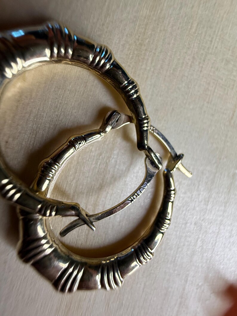 May include: Gold-toned hoop earrings with a bamboo-like design. The earrings have a textured surface and a hinged closure. The metal is stamped with "AU 10K". These earrings are a stylish accessory.