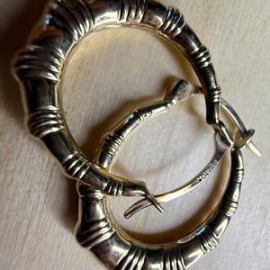 May include: A pair of gold-toned hoop earrings with a bamboo-inspired design. The earrings have a textured surface and a hinged closure. The metal is stamped with "10K".