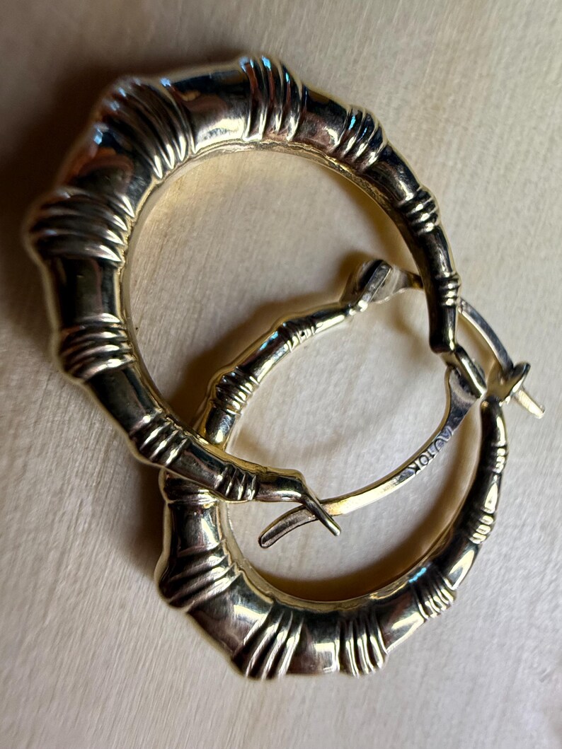 May include: A pair of gold-toned hoop earrings with a bamboo-inspired design. The earrings have a textured surface and a hinged closure. The metal is stamped with "10K".