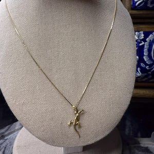 May include: A gold-colored necklace featuring a lizard pendant. The lizard is detailed with outstretched limbs and a curved tail. The necklace is displayed on a beige mannequin.