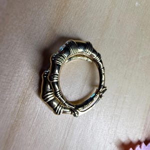 May include: A gold-colored ring with a bamboo design. The ring is circular, with the bamboo stalks forming the band. The ring is on a light brown surface.
