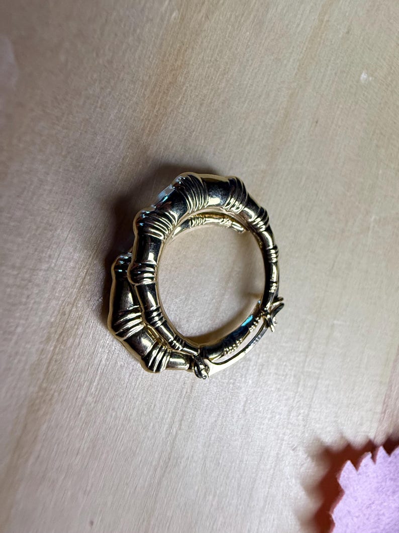 May include: A gold-colored ring with a bamboo design. The ring is circular, with the bamboo stalks forming the band. The ring is on a light brown surface.