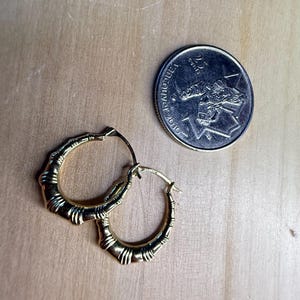 May include: A pair of gold-colored hoop earrings with a bamboo-like design. The earrings are next to a Canadian quarter coin. The earrings are on a light brown wooden surface.