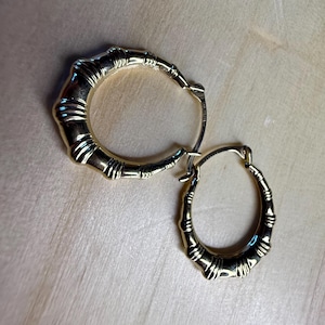 May include: A pair of gold-colored hoop earrings with a bamboo-like design. The earrings are oval-shaped and feature detailed ridges and segments, creating a textured appearance. The earrings are displayed on a light-colored wooden surface.