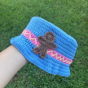 May include: A handmade blue crochet bucket hat with a pink and yellow band and a brown gingerbread man applique. The hat is displayed against a green grassy background.